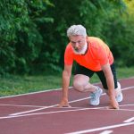 Full body of confident focused senior male ready for sprint on racetrack in stadium near green bushy plants